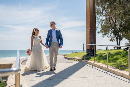 A newly wed couple walking hand in hand down a path in front of a beach.