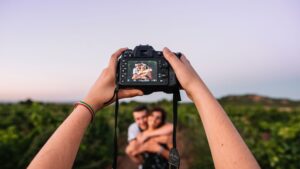 Photographer holding up camera in focus of a couple hugging in field.