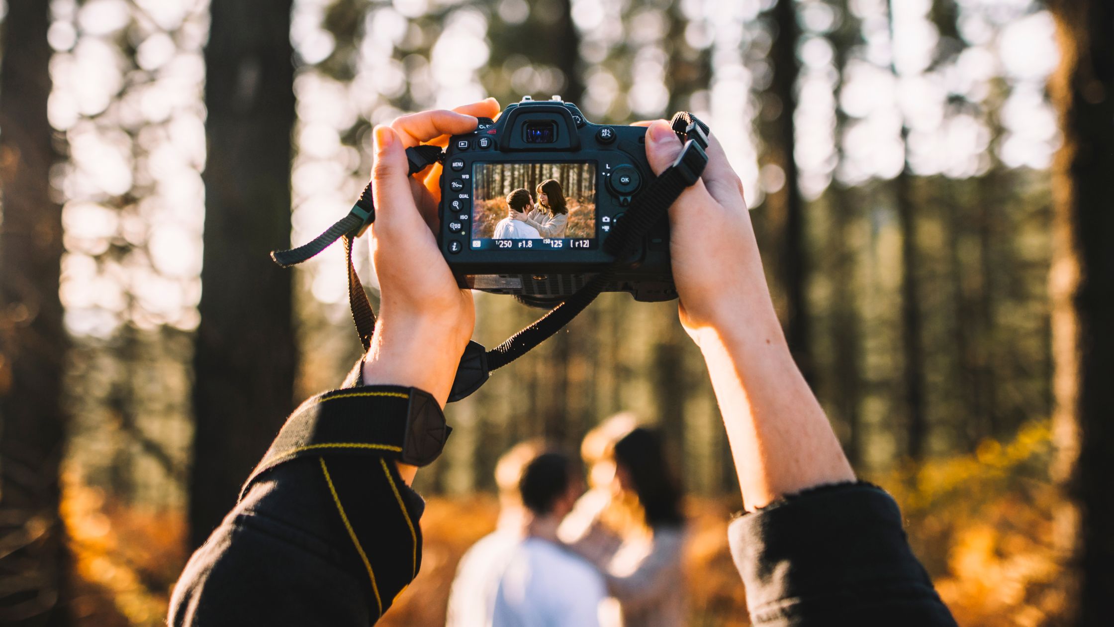 Photographer holding up camera in focus of a couple hugging in field.